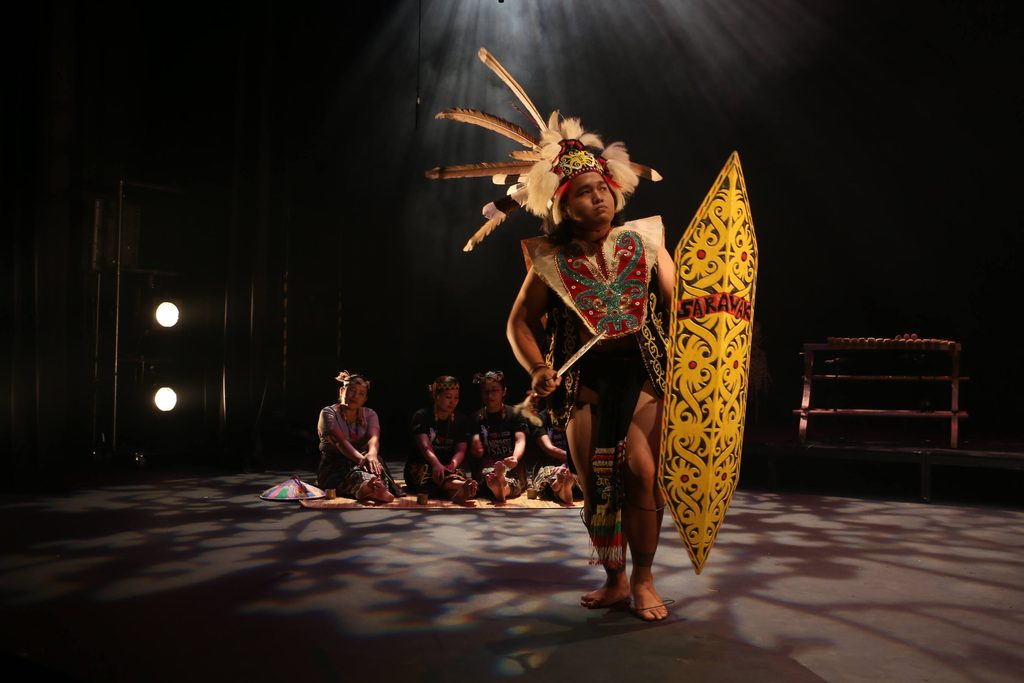 A cultural bearer performing a traditional Kayan warrior dance with a shield during a live cultural production