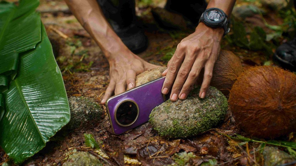 Hands resting a purple HONOR X9e Pro smartphone on a mossy rock next to whole coconuts on the forest ground
