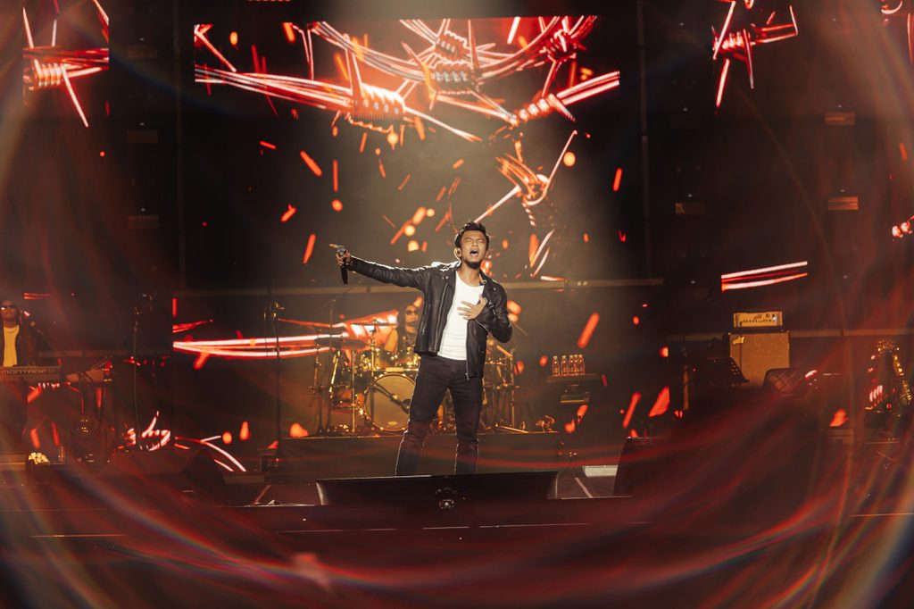 Faizal Tahir berjaket kulit menyanyi di tengah pentas besar berlatarbelakangkan grafik visual kawat berduri dan percikan api